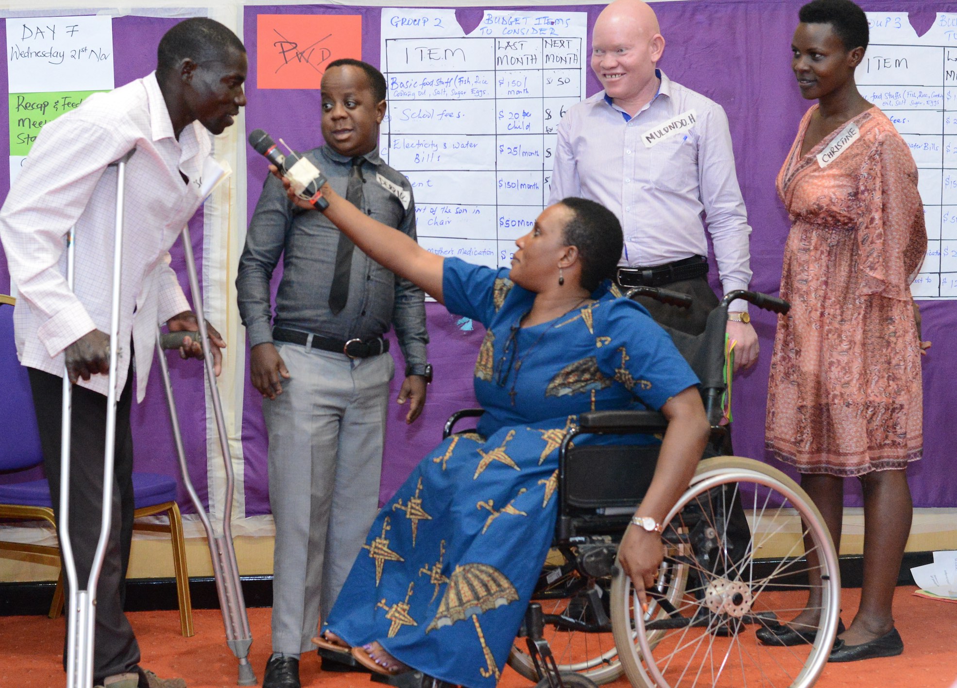 Participants at a GDS meeting. A lady wheelchair user holds a microphone to a man who is speaking. Three other participants stand next to them smiling. 