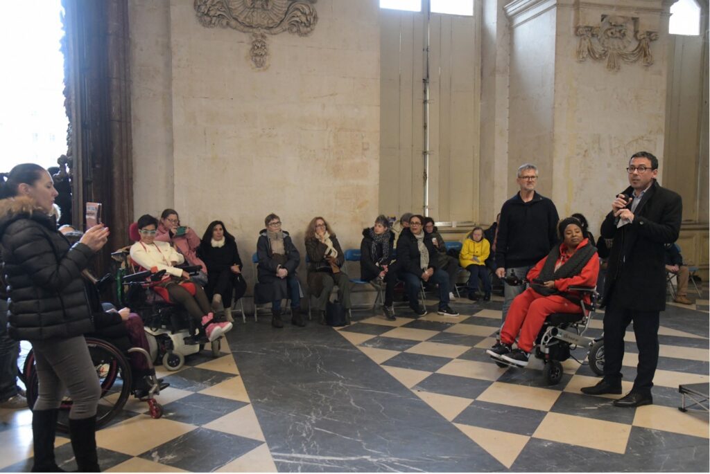 A speaker addresses an audience gathered in a historic hall during an inclusive event, with people of diverse ages and abilities seated and using mobility devices, including wheelchairs and powered wheelchairs. The gathering is part of the City of Lyon's Disability Fortnight, celebrating the International Day of Persons with Disabilities and the city's inclusive policies. Photo credit: Muriel Chaulet.