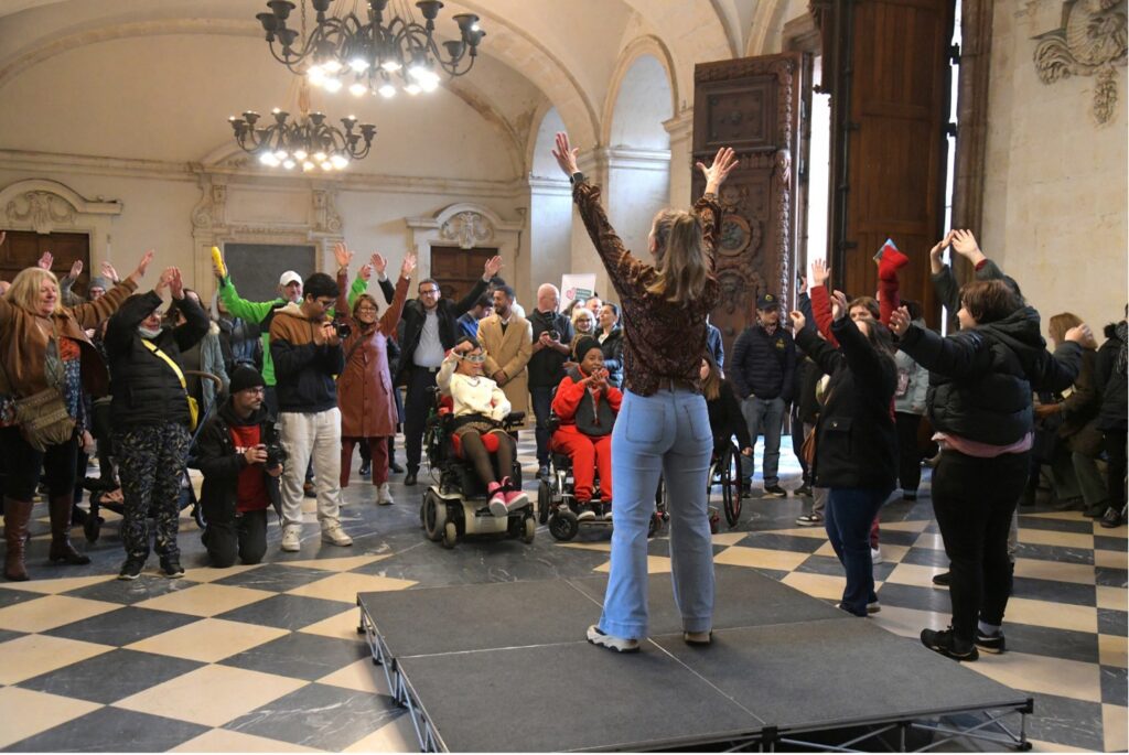 People of diverse ages and abilities gather in a historic hall, raising their hands together during an  activity led by a facilitator standing on a small stage. The event celebrates the International Day of Persons with Disabilities as part of the City of Lyon's Disability Fortnight. Photo credit: Muriel Chaulet.