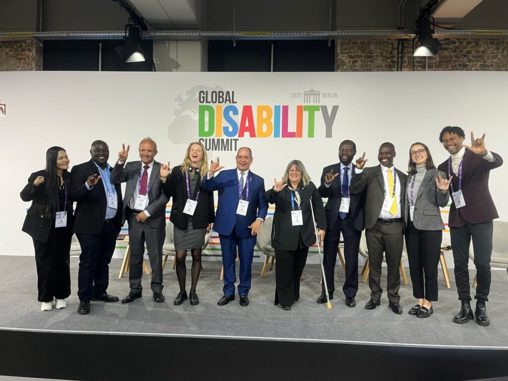 Nine people of different backgrounds, ages and disabilities pose on stage at the 2025 Global Disability Summit in Berlin, several using the ASL “I love you” sign.